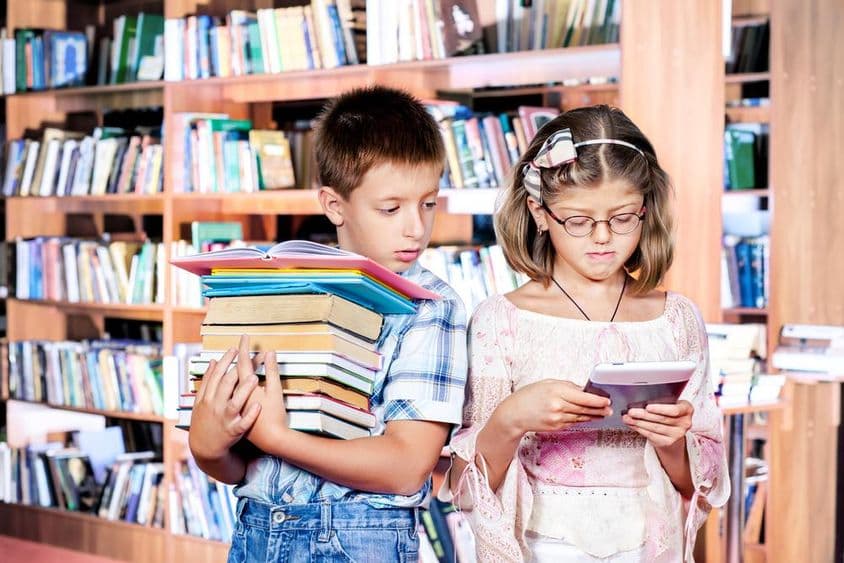لائبریری میں دو بچے، ایک ای بک پڑھ رہا ہے، دوسرا ہاتھوں میں کئی کتابیں لیے ہوئے ہے۔