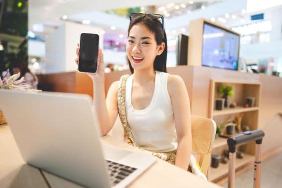 نسل زیڈ کی لڑکی کام کر رہی ہے، فون پکڑے ہوئے ہے، اس کے سامنے ایک نوٹ بک ہے اور اس کے پاس ایک سفری سوٹ کیس کا ہینڈل ہے۔