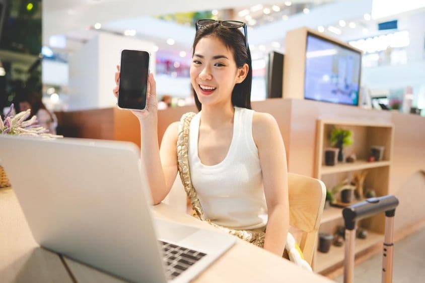 نسل زیڈ کی لڑکی کام کر رہی ہے، فون پکڑے ہوئے ہے، اس کے سامنے ایک نوٹ بک ہے اور اس کے پاس ایک سفری سوٹ کیس کا ہینڈل ہے۔