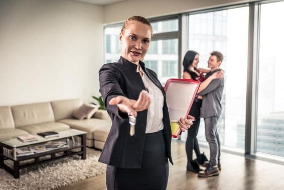 ایک خاتون رئیل اسٹیٹ بروکر ہاتھ ہلاتے ہوئے، پس منظر میں ایک جوڑا گلے لگا رہا ہے۔