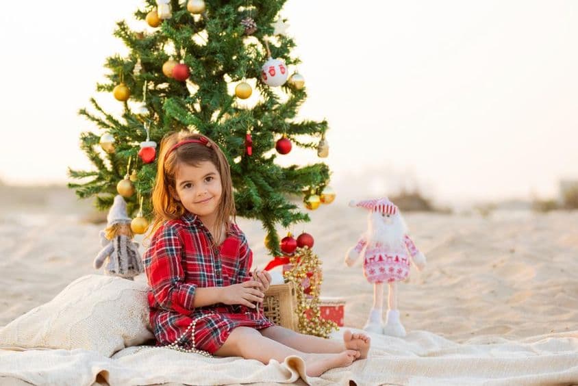 کرسمس درخت کے نیچے ساحل پر بیٹھی خوش باش ہنس رہی چھوٹی لڑکی۔