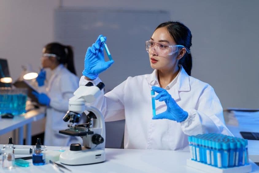 لیب کوٹ اور دستانے میں ایشیائی خاتون سائنسدان دو ٹیسٹ ٹیوبز کا موازنہ کرتے ہوئے