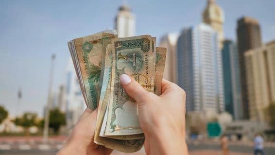 ایک لڑکی یو اے ای کرنسی پکڑے ہوئے، پس منظر میں دبئی کے بلند و بالا عمارات۔