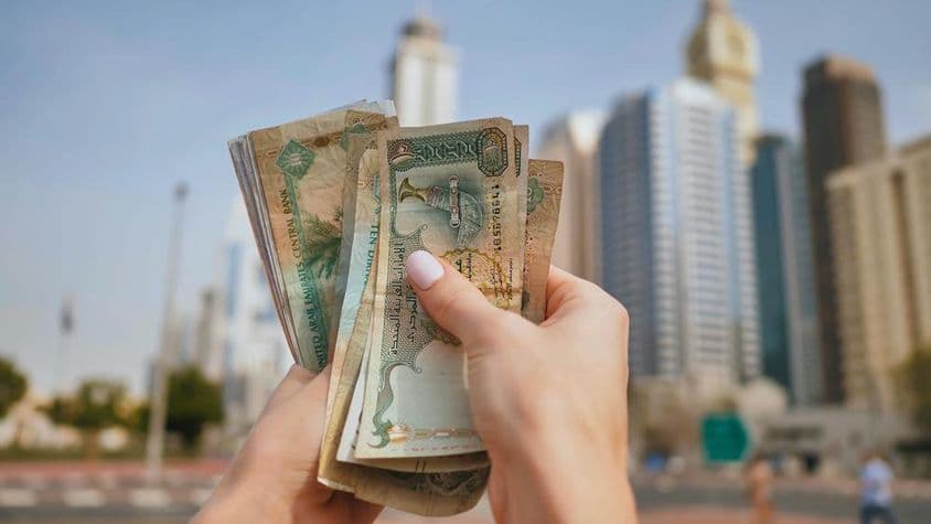 ایک لڑکی یو اے ای کرنسی پکڑے ہوئے، پس منظر میں دبئی کے بلند و بالا عمارات۔