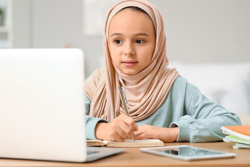 گھر میں حجاب پہنے ہوئے ایک چھوٹی مسلم لڑکی ہوم ورک کرتی ہوئی۔